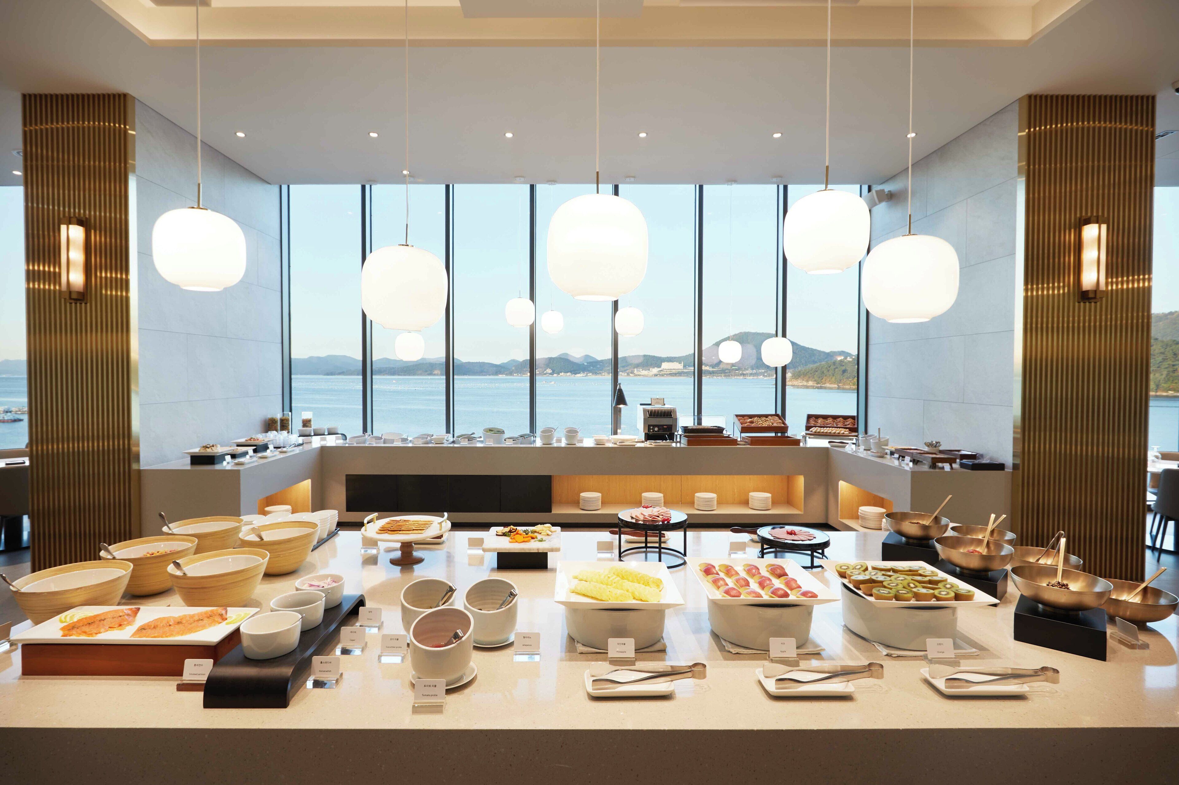 Montaje de bufé con diferentes alimentos y utensilios, ventanas amplias con vistas al mar y lámparas colgantes.