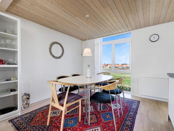 A room with a round table, chairs, a lamp, a bookshelf, a decorative wreath, and a window with a view.