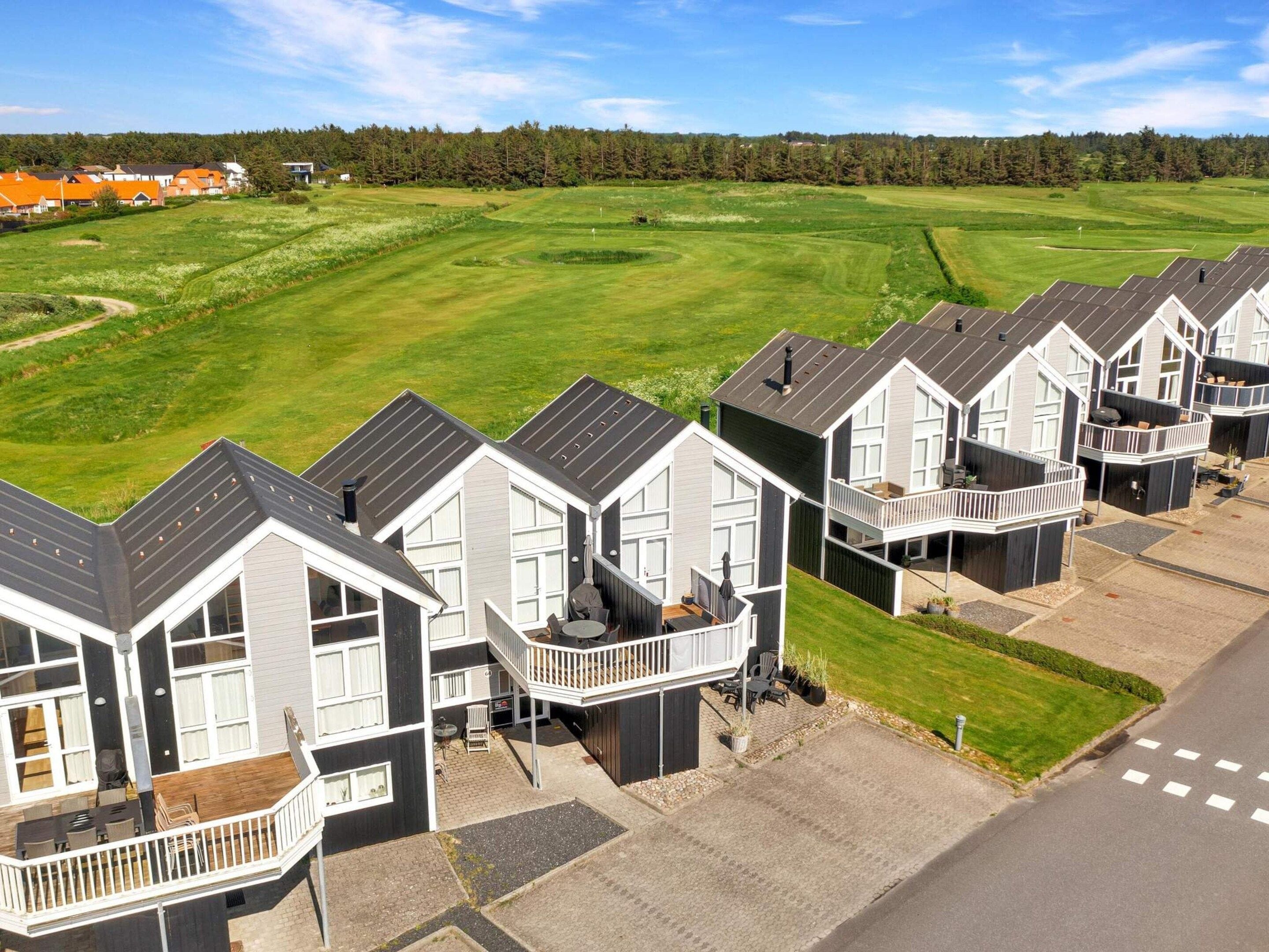 A row of modern, two-story houses with balconies, set against a backdrop of a well-maintained golf course and a forested area.