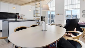 A modern kitchen with a central island, a dining area with a round table, and a view of the outdoors through large windows. - 5 Star Holiday Home in Lokken (Lokken)