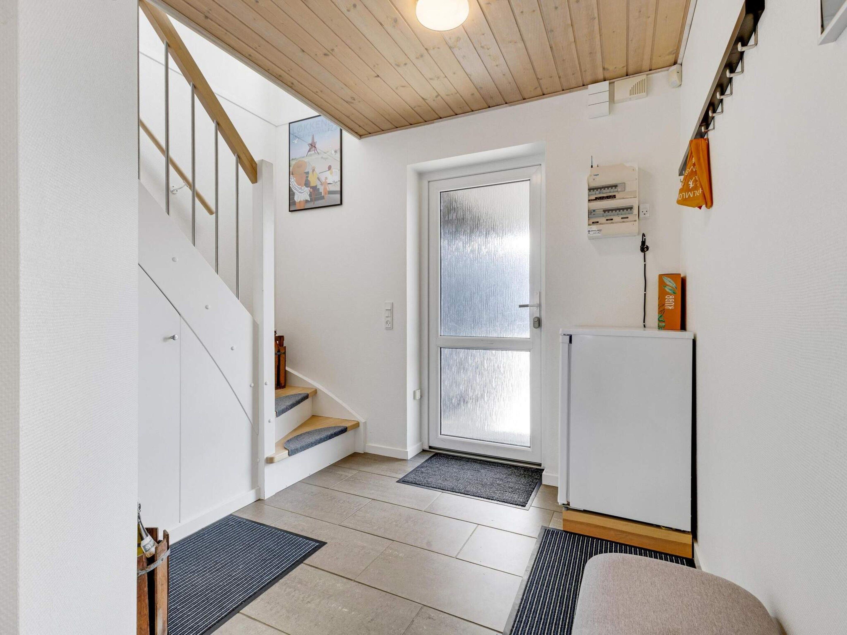 A hallway with a wooden staircase, a door with frosted glass, a white wall, and a small refrigerator.