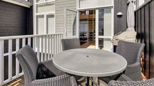 A covered outdoor patio with a round table and wicker chairs, a grill, and a view of the interior through large windows. - 5 Star Holiday Home in Lokken (Lokken)