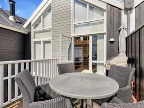 An outdoor patio with a round table and wicker chairs, a white railing, and a building with large windows and a chimney.