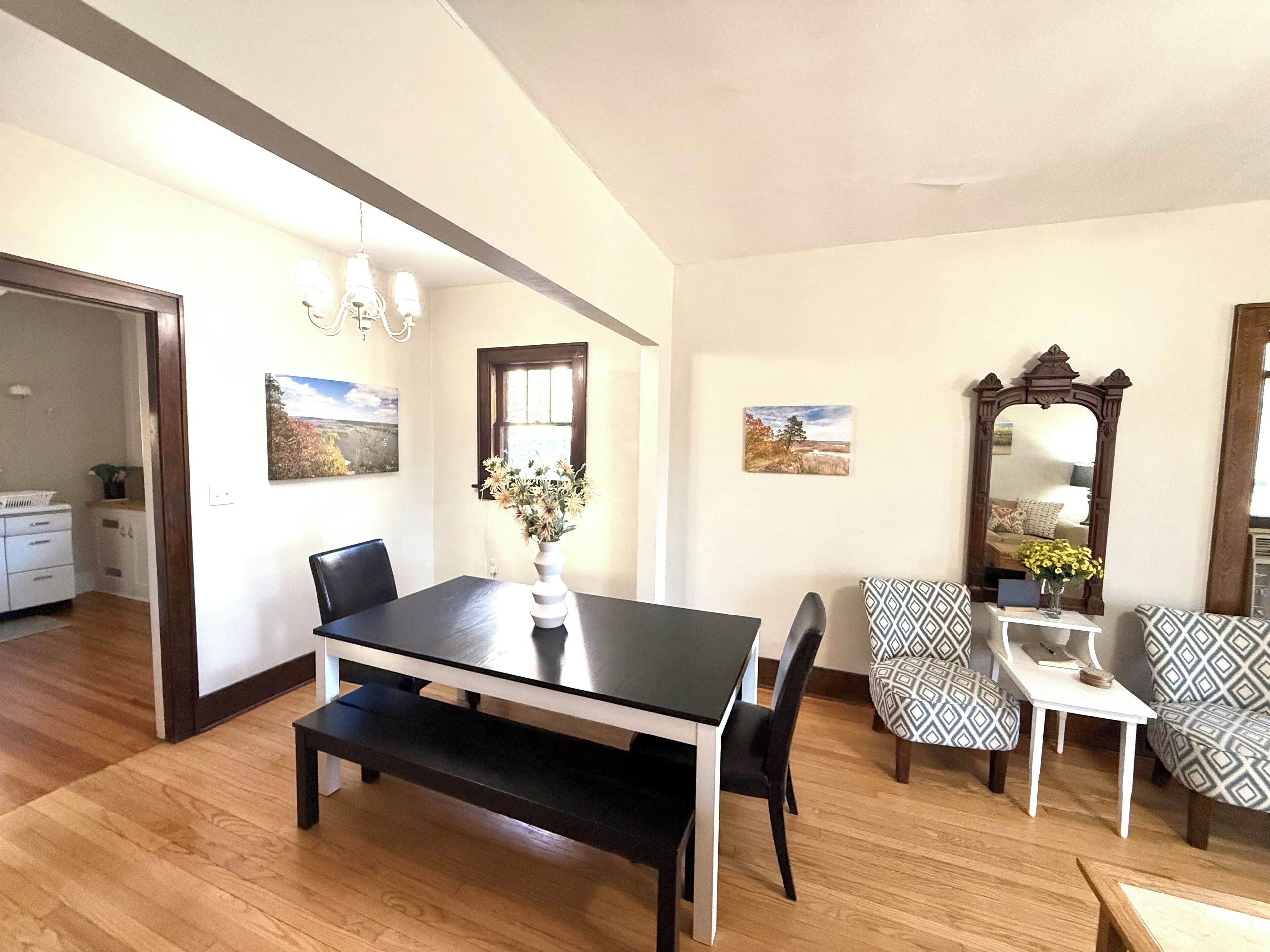 A dining area with a black table, white bench, patterned chairs, a vase with flowers, and a large mirror.