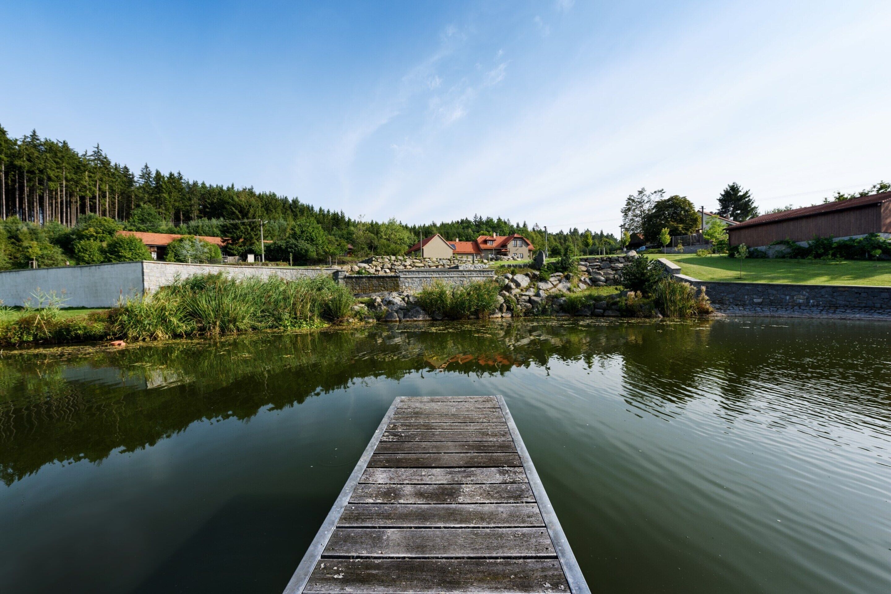 Une jetée en bois s’étendant sur un lac paisible, avec un mur de pierres et de la verdure en arrière-plan.