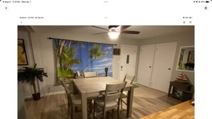 A dining area with a wooden table and chairs, a ceiling fan, and a view of the outdoors through a window with a tropical scene backdrop.