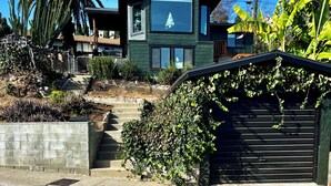 Une maison moderne à deux étages, dotée d’une grande fenêtre au deuxième étage, entourée d’une végétation luxuriante et accessible par un petit escalier menant à l’entrée.