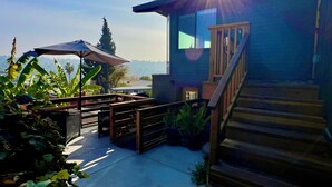 Une maison en bois avec un escalier menant à un balcon, entourée de plantes en pot et d’un grand parasol.