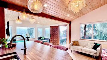 Een moderne keuken met een houten aanrecht en een spoelbak, een eethoek met een grote tafel en roze bekleden stoelen, en een woonkamer met een witte bank en een groot raam met uitzicht op groen.