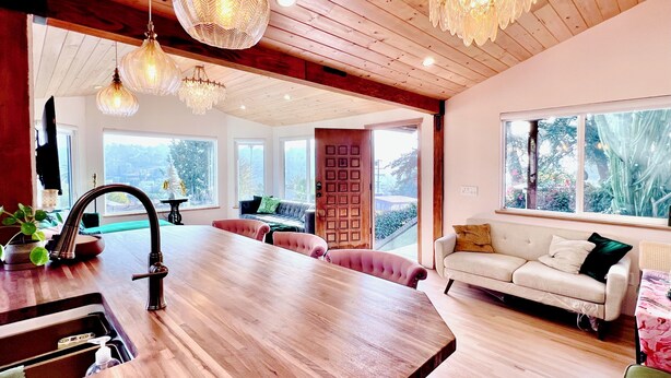 Une cuisine moderne avec un plan de travail en bois et un évier, un coin repas avec une grande table et des chaises à revêtement rose, et un salon avec un canapé blanc et une grande fenêtre donnant sur un paysage verdoyant.