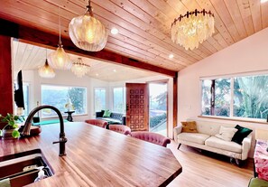 Una cocina moderna con encimera de madera y un lavamanos, un comedor con una mesa grande y sillas tapizadas en rosa, y una sala de estar con un sofá blanco y un ventanal que ofrece vistas a zonas verdes.