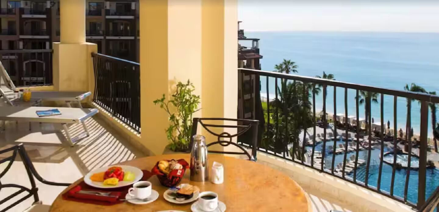 Un balcón con una mesa dispuesta para el desayuno, vista al océano y zona de piscina.