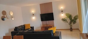 A modern living room with a black sofa, a flat-screen TV, wall-mounted lights, and a potted plant.