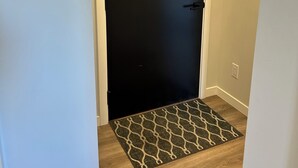 A closed door with a black surface, a doormat with a geometric pattern, and a light switch on the wall. - Quick walk to Lakeside Boardwalk and Downtown shops & restaurants! (Kelowna)