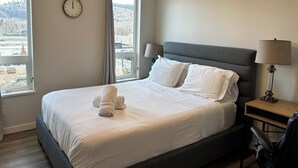 A hotel room with a bed, two bedside lamps, a clock, and a view of the outside. - Quick walk to Lakeside Boardwalk and Downtown shops & restaurants! (Kelowna)