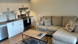 A modern kitchen with white cabinets, a built-in oven, and a stainless steel range hood. There is a beige sofa with patterned cushions and a small wooden coffee table. - Quick walk to Lakeside Boardwalk and Downtown shops & restaurants! (Kelowna)