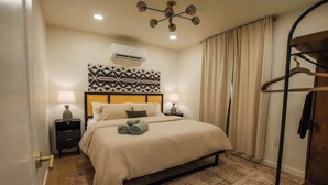 Quarto de hotel bem arrumado, com uma cama, duas mesas de cabeceira, luminária pendente e ar-condicionado fixado na parede.