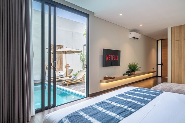 A modern hotel room with a large bed, a TV mounted on the wall, and a sliding glass door leading to an outdoor pool area.