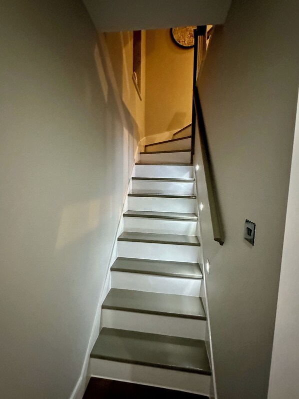 Eine Treppe mit weißen Stufen und einem schwarzen Geländer, die zu einer Zwischenebene mit Lichtschalter und Wandleuchte führt.