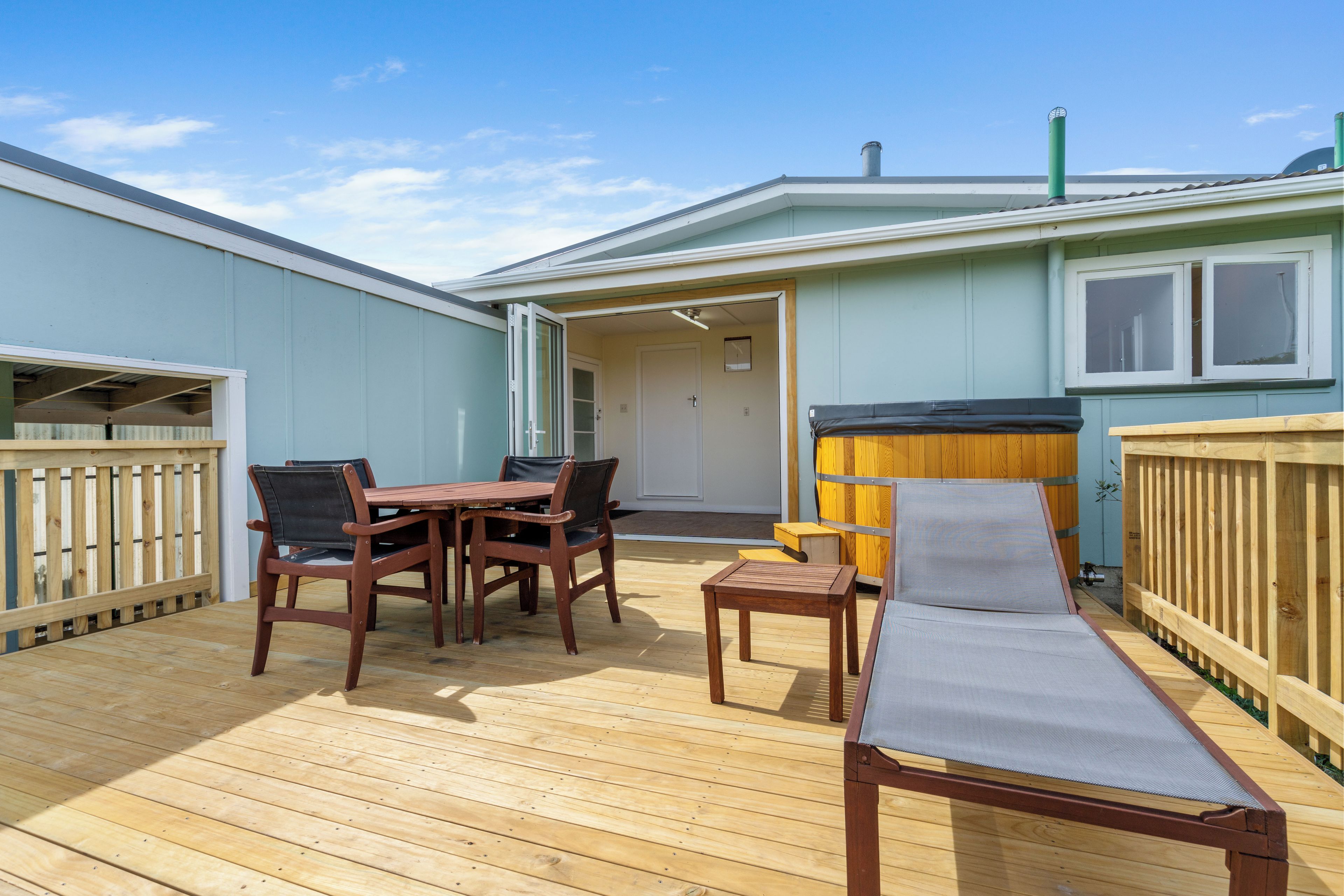 A covered outdoor deck with a table and chairs, a lounge chair, and a wooden fence.