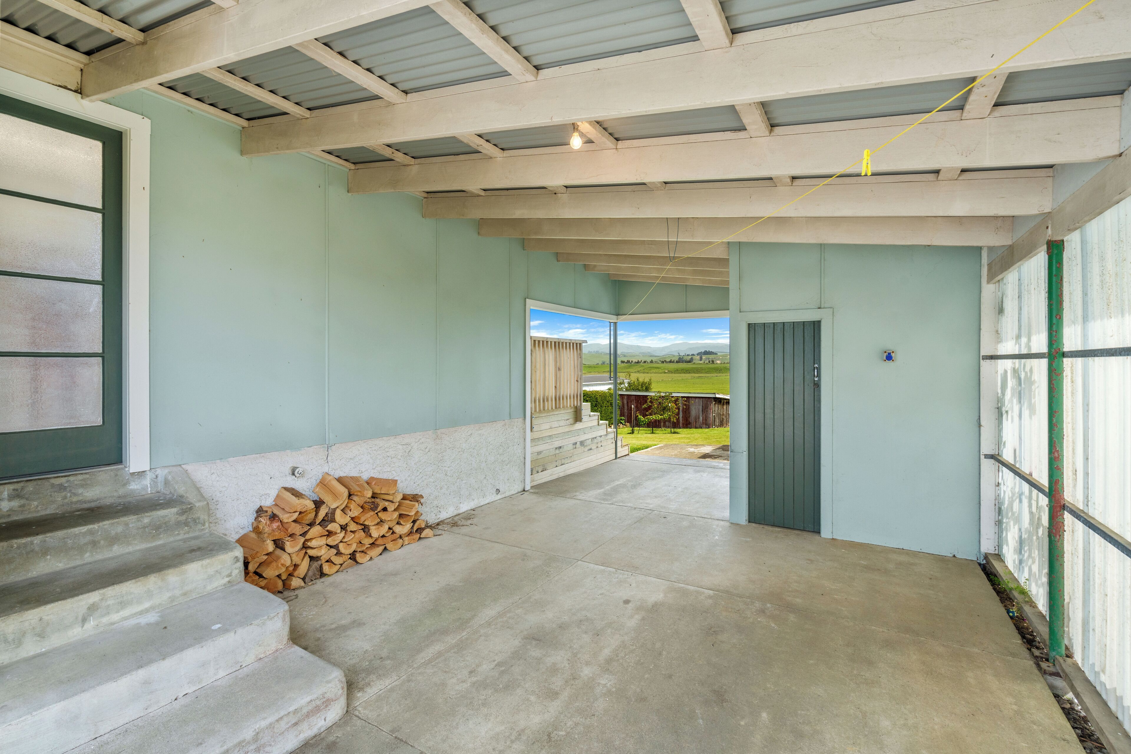 A covered outdoor area with concrete flooring, a stack of firewood, and a view of a green landscape through an open door.