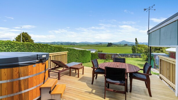 Una terraza de madera con un jacuzzi, una mesa y sillas, con vistas a un paisaje verde con colinas.