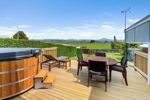 Une terrasse en bois avec un jacuzzi, une table et des chaises, donnant sur un paysage verdoyant et vallonné.