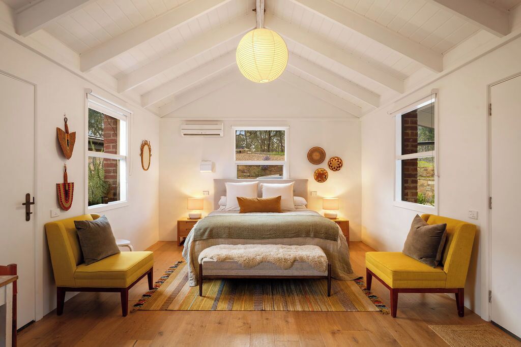 A bedroom with a bed, two yellow chairs, a hanging light fixture, and a window with a view.