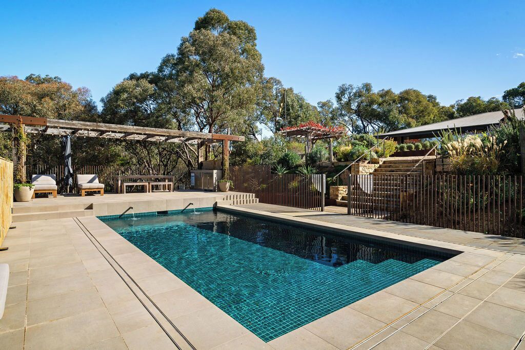 Una piscina rectangular con agua azul cristalina, rodeada por un patio de baldosas. Hay sillas de descanso y una pérgola con techo.