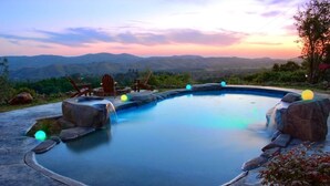 Une piscine avec vue sur les montagnes au coucher du soleil, entourée de rochers et de mobilier d’extérieur.
