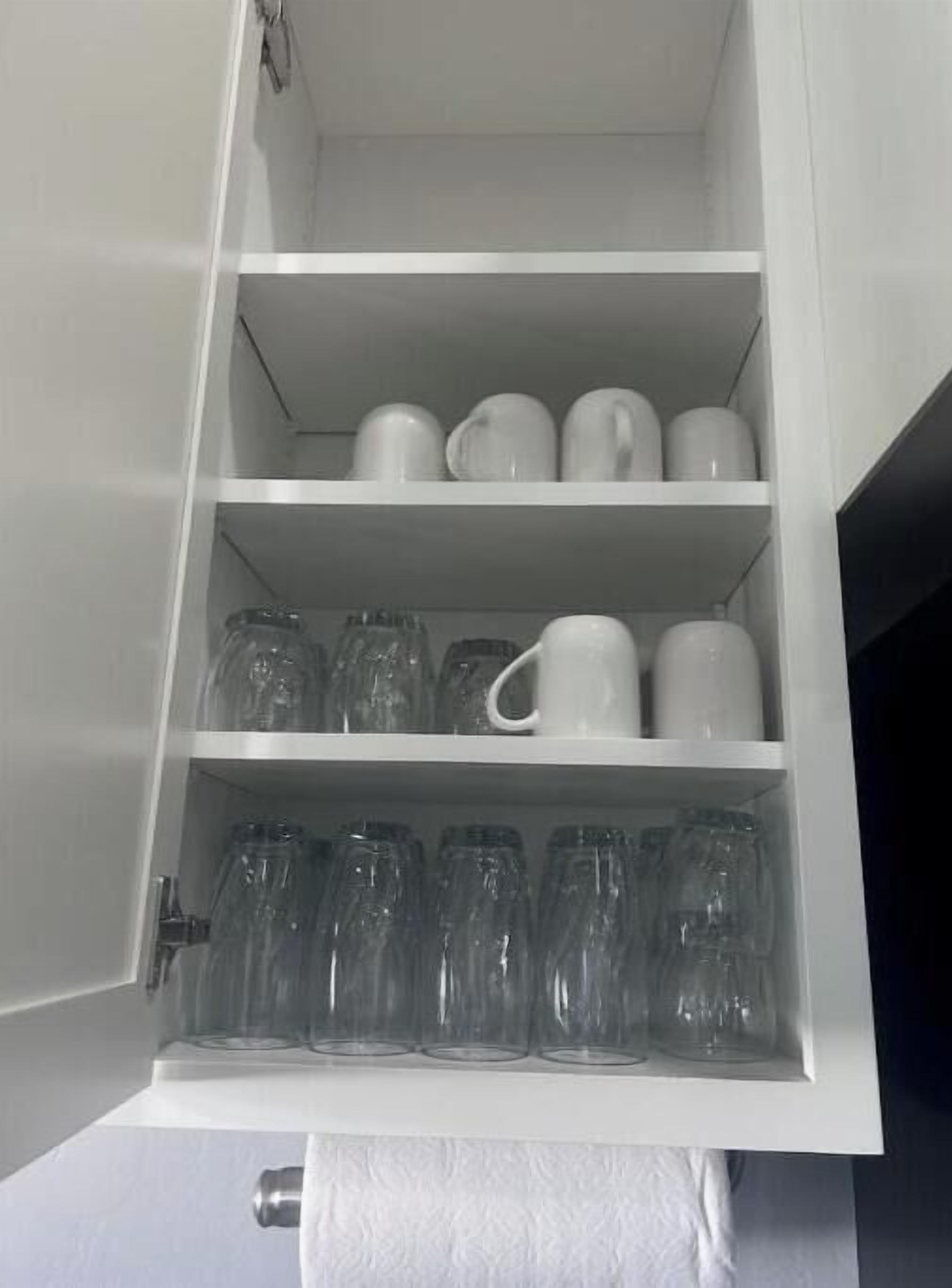 A white cabinet with shelves containing clear glass bottles and white mugs.