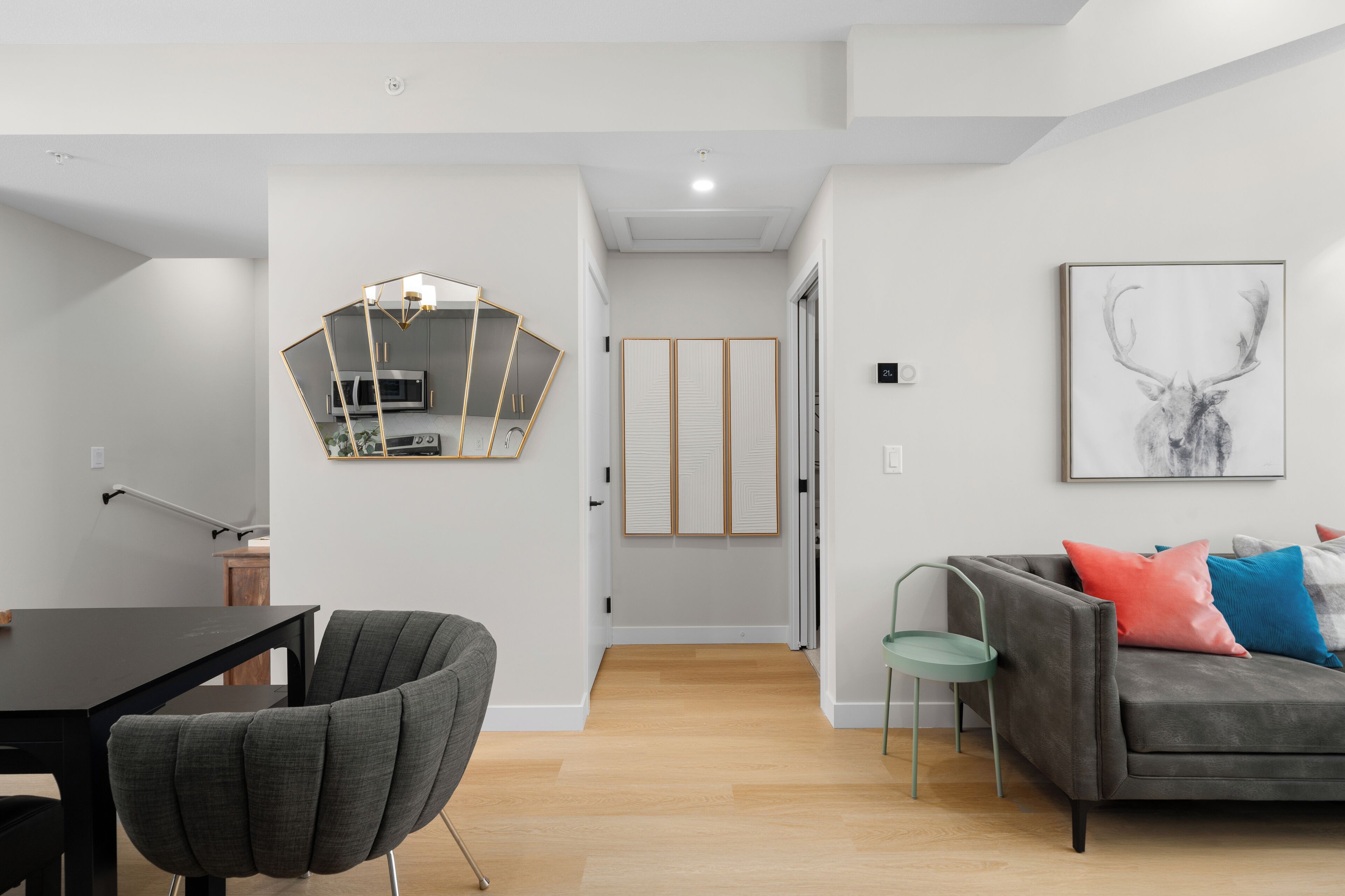 A modern living room with a grey sofa, a dining table with chairs, a kitchen area with a microwave, and a hallway leading to a door.