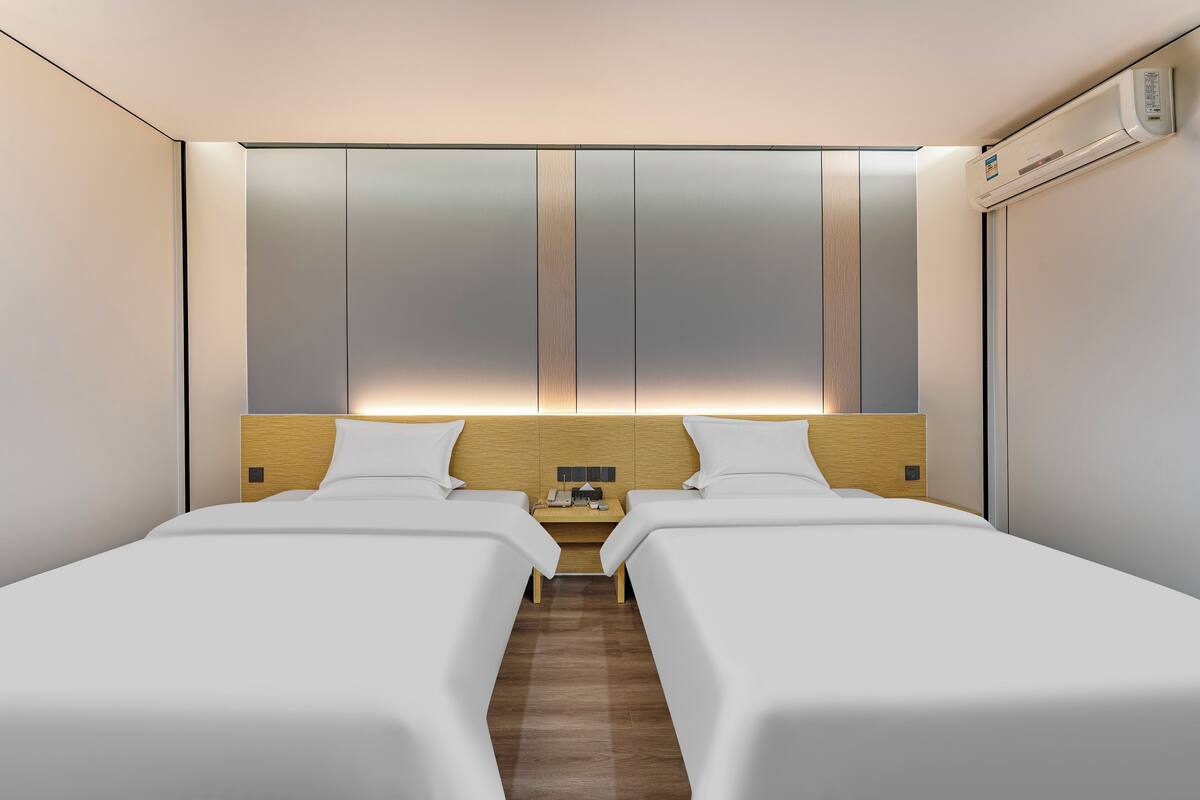 Une chambre d’hôtel avec deux lits simples, une tête de lit en bois, une table de chevet avec un téléphone et un appareil de climatisation.