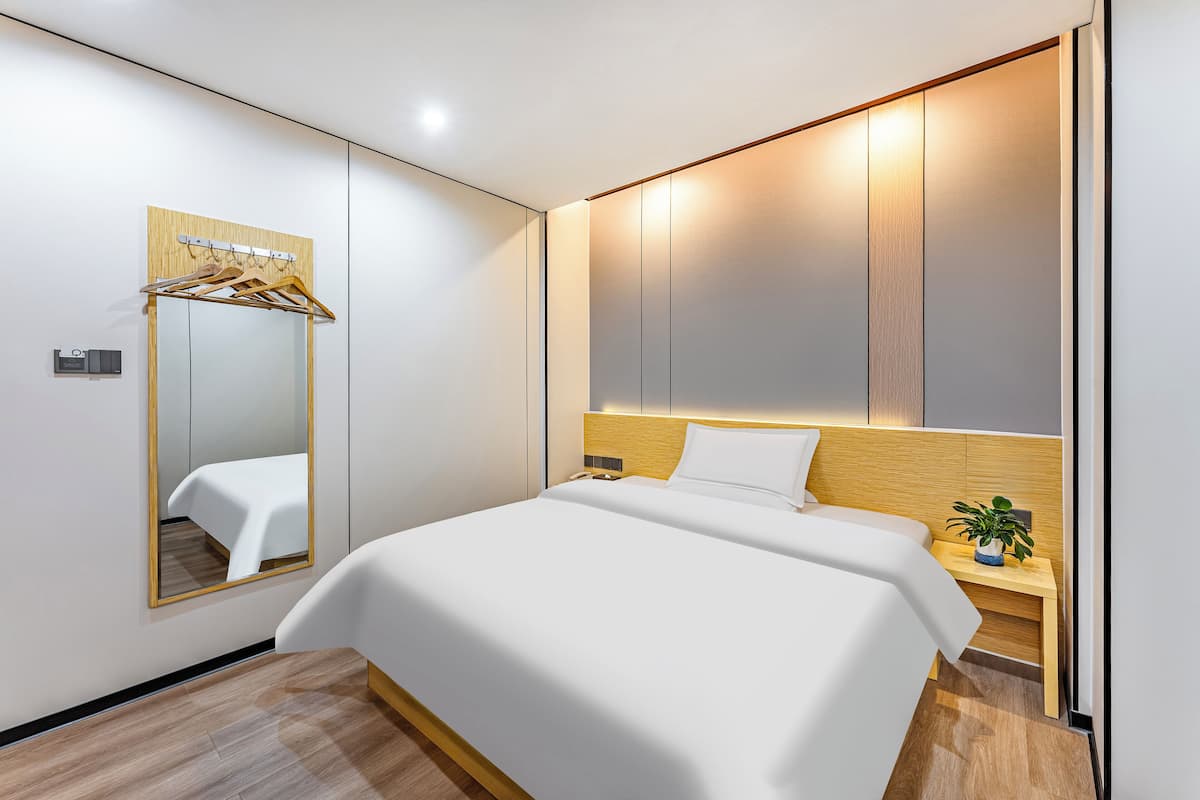 Une chambre d’hôtel moderne avec un grand lit, une tête de lit en bois, une table de chevet agrémentée d’une plante et un miroir avec un porte-manteau en bois.