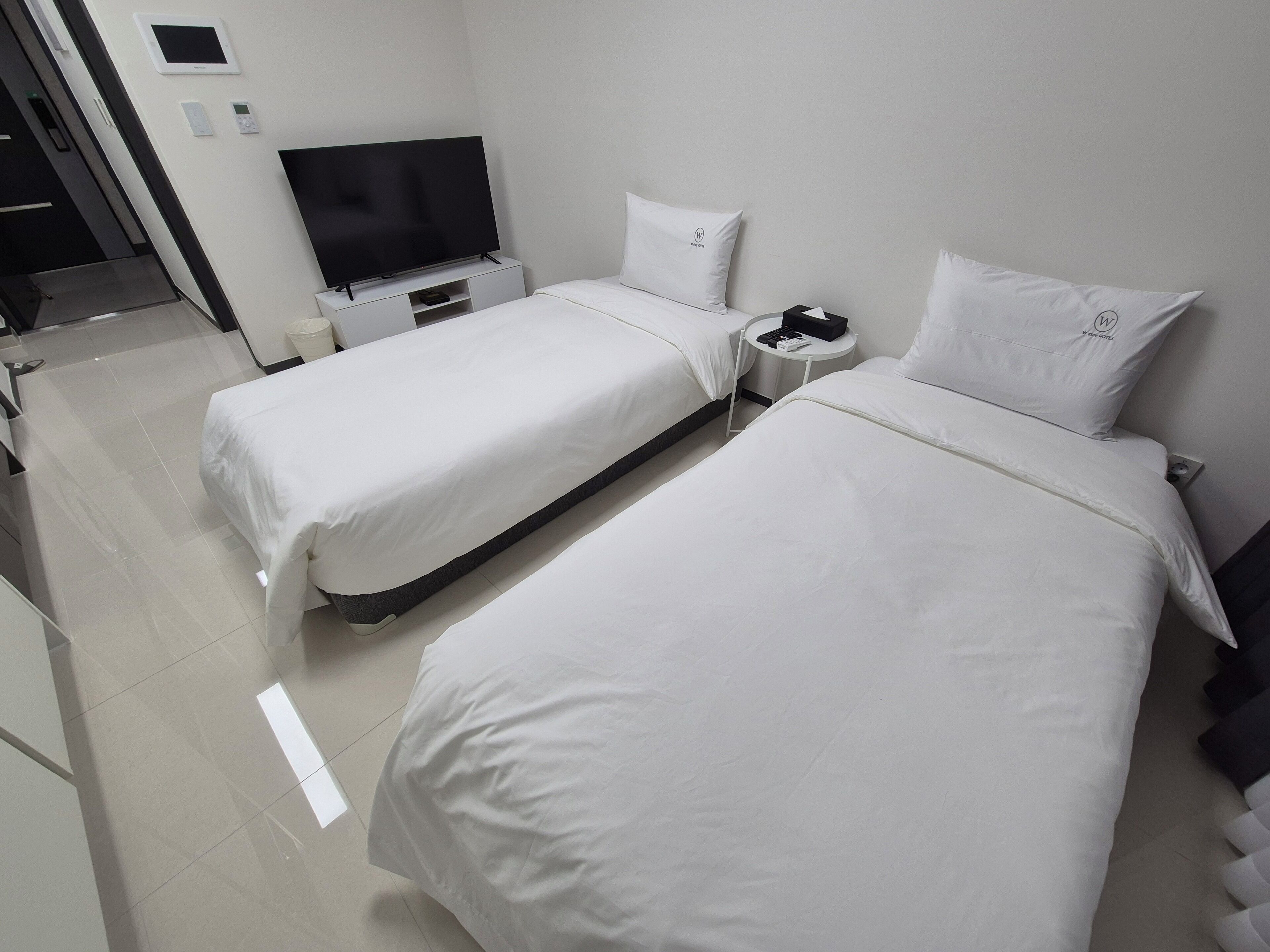 A hotel room with two beds, a TV, a small table, and a wall-mounted control panel.