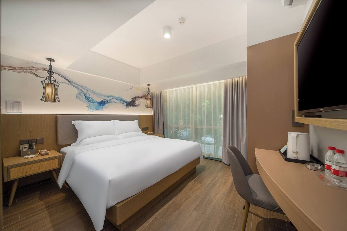A hotel room with a bed, bedside table, chair, TV, and a window with curtains.