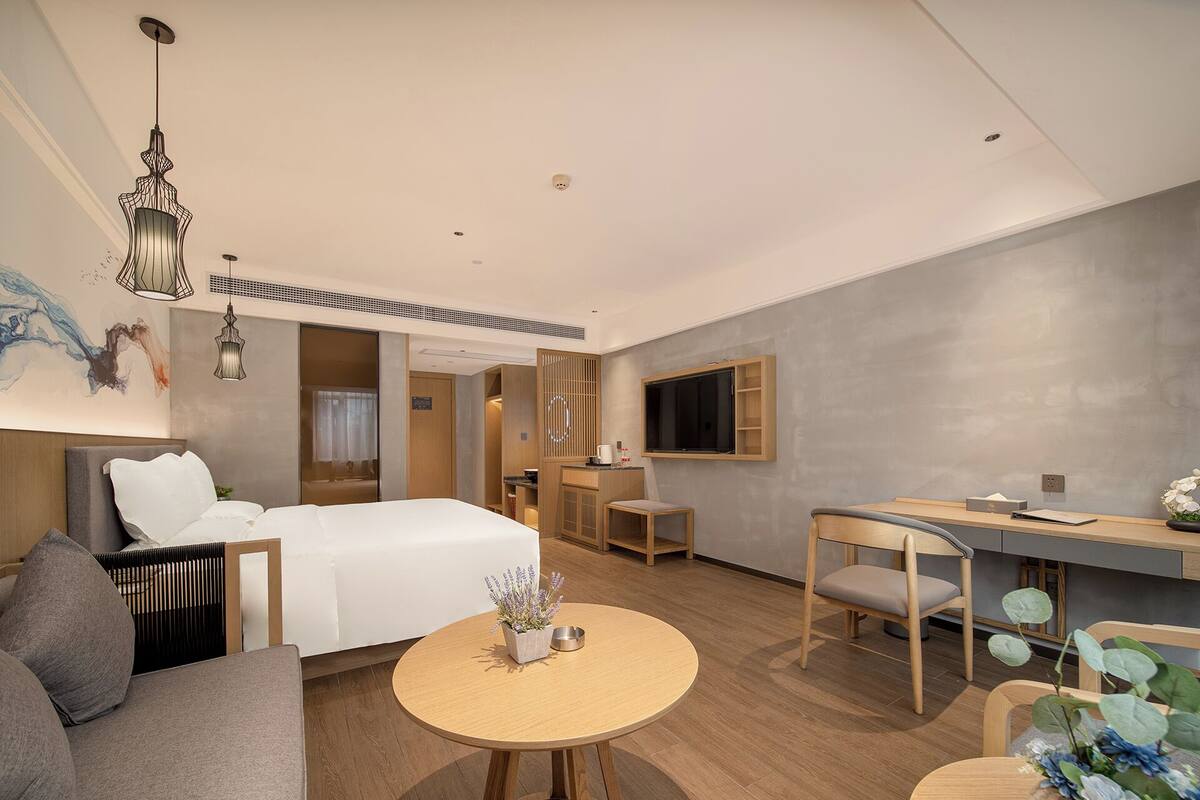 A modern hotel room with a bed, a sofa, a dining table, a TV, and a small kitchen area.