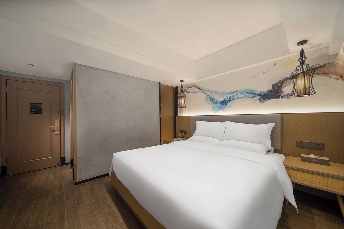 A hotel room with a large bed, a wooden nightstand, a wall-mounted lamp, and a door with a sign.
