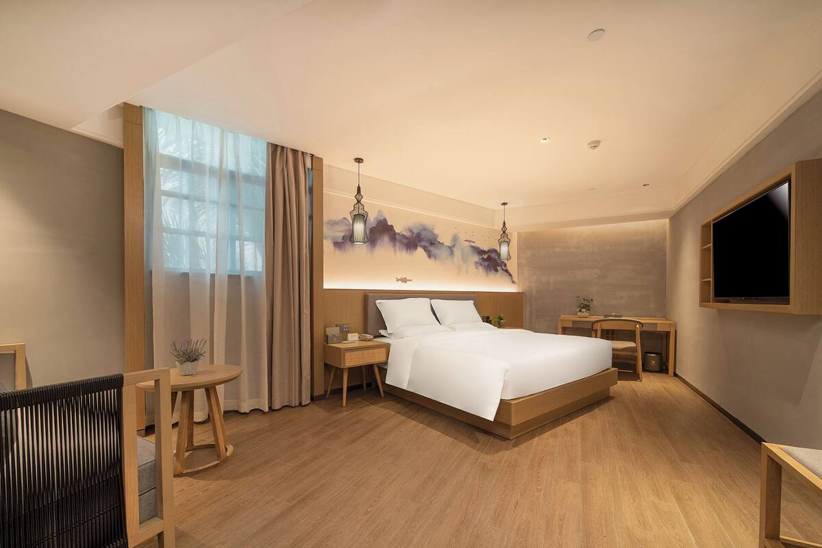 A modern hotel room with a large bed, a desk, a chair, a TV, and a window with curtains.