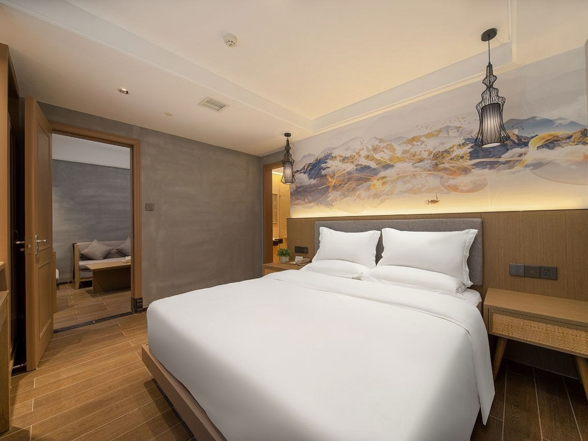 A modern hotel room with a large bed, a wooden nightstand, a wall-mounted lamp, and a painting of mountains.
