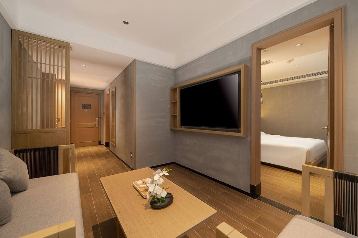 A hotel room with a bed, a TV mounted on the wall, a wooden table with a vase of flowers, and a sliding door leading to another room.