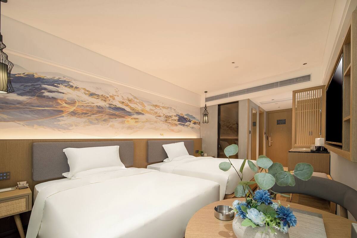 A hotel room with two beds, a large abstract mural on the wall, a round wooden table with a vase of flowers, and a television on the right.