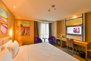 Een hotelkamer met een groot bed, een bureau met een computer, paarse stoelen en een schilderij van een veld.