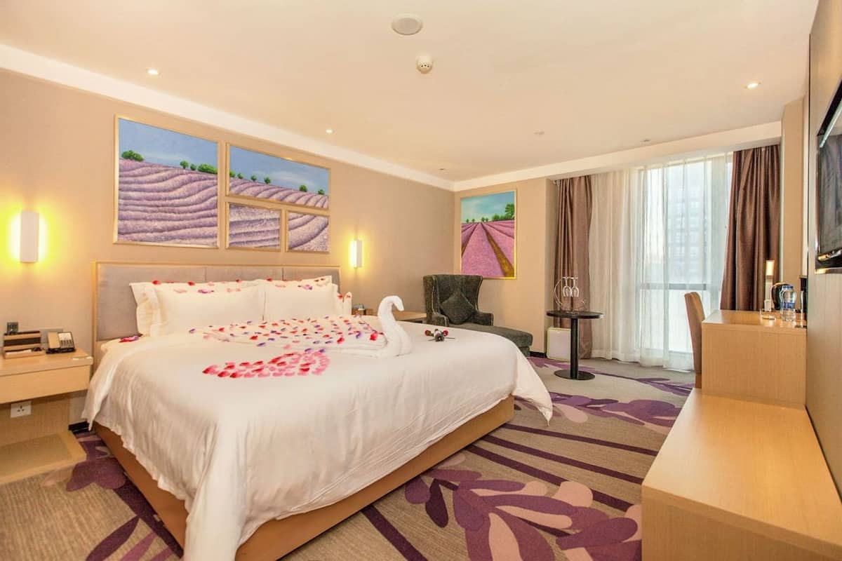 A hotel room with a large bed, a desk, a chair, and a TV. There are paintings on the wall and a window with curtains.