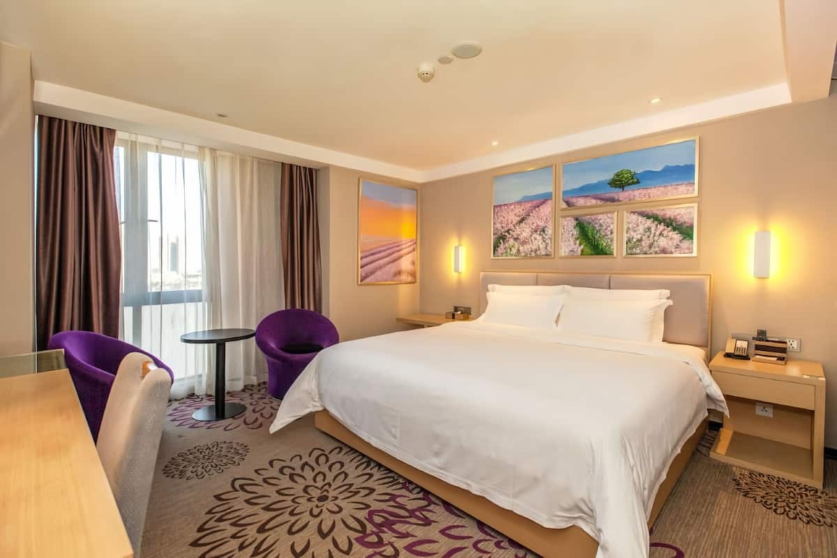 A hotel room with a large bed, a small table, purple chairs, and a view of a field through the window.