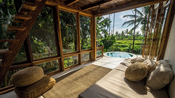 Une pièce dotée d’une grande fenêtre donnant sur une piscine et une végétation luxuriante. On y trouve des tapis et des coussins en fibres naturelles disposés sur un lit.