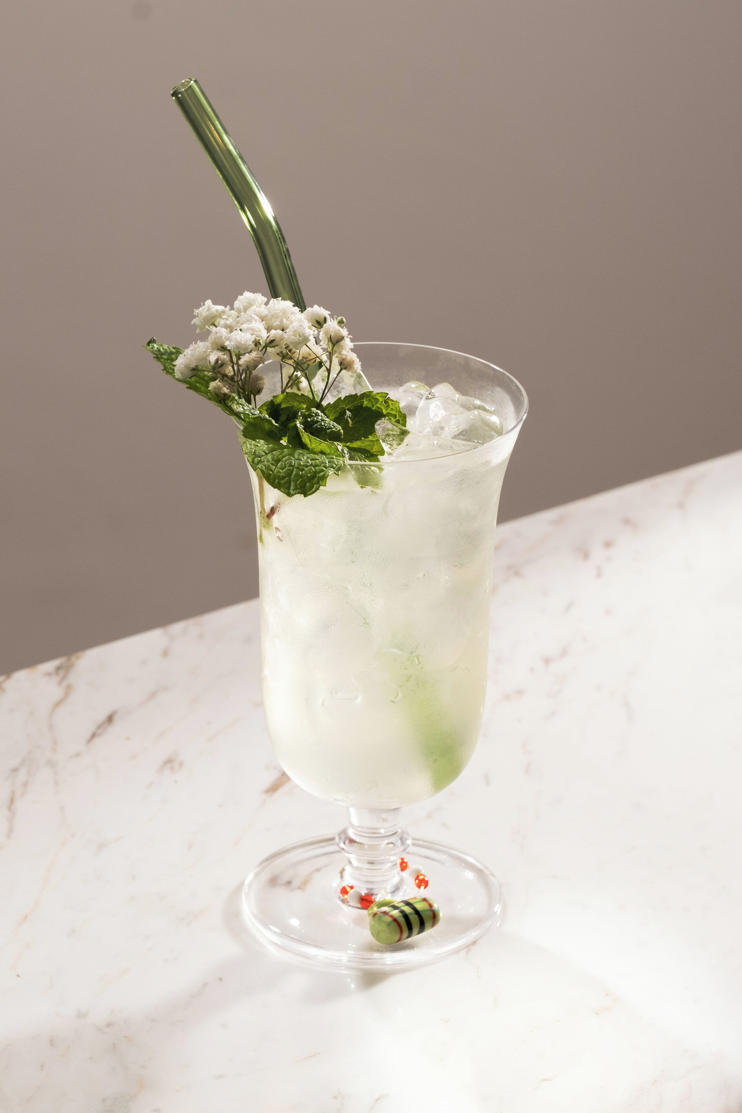 A tall glass with a green stem, filled with a clear liquid and ice, garnished with mint leaves and a small flower arrangement.