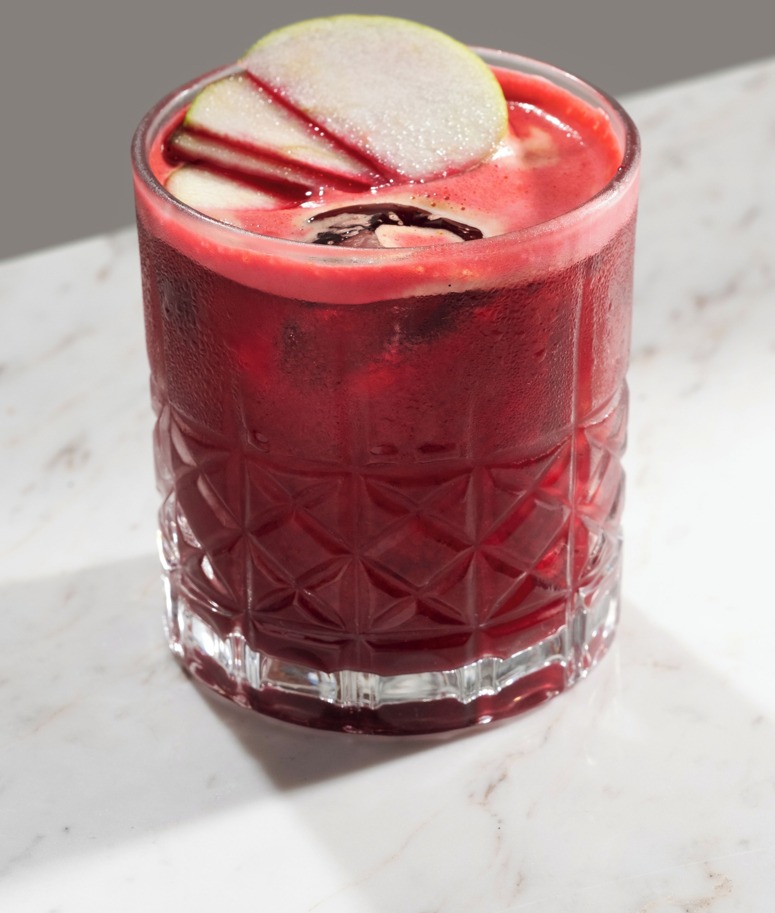 A glass with a textured pattern and a red beverage, possibly a cocktail, with a slice of lime on the rim.