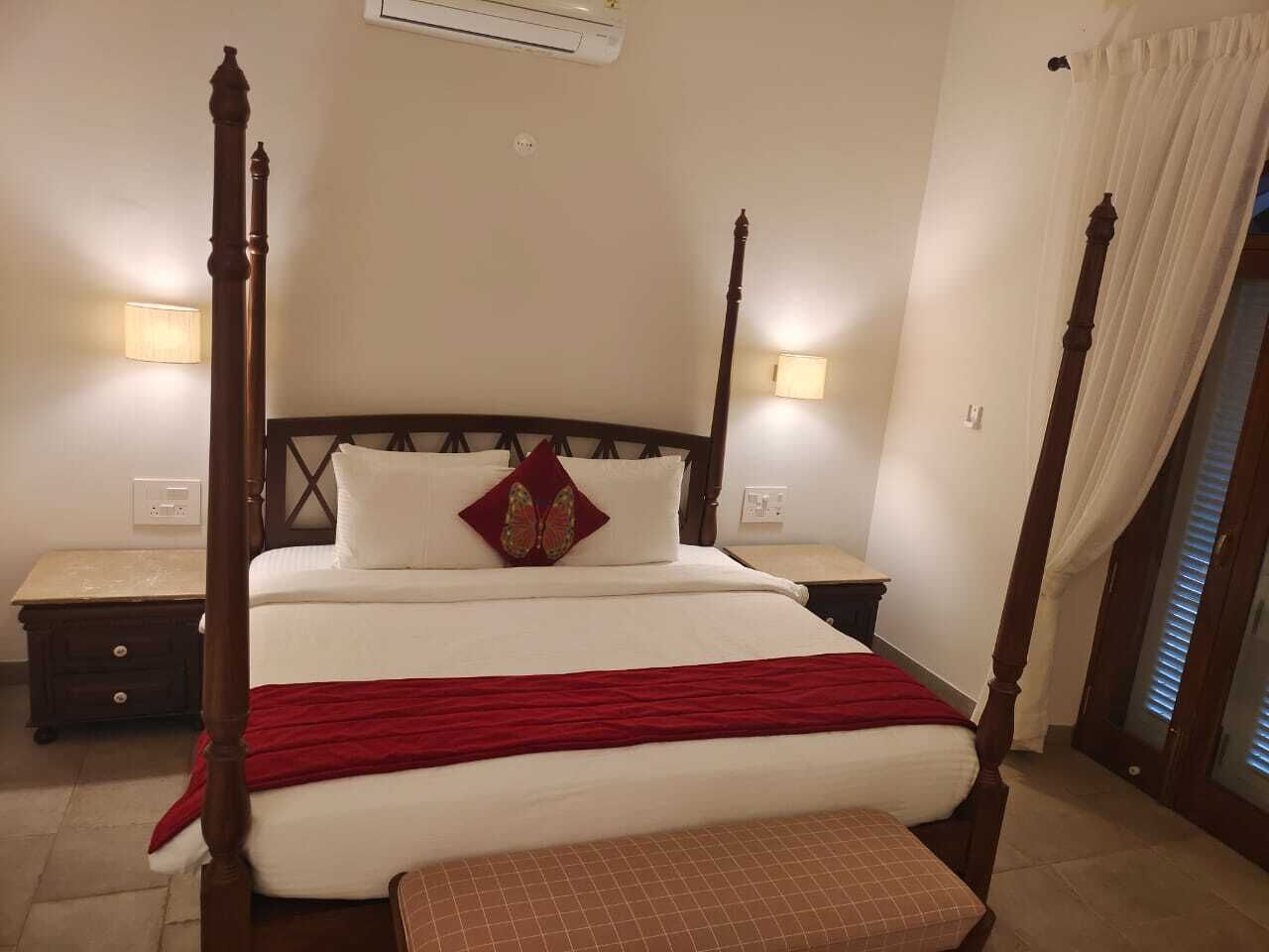 A four-poster bed with a red throw blanket, a wooden nightstand, and a wall-mounted lamp.