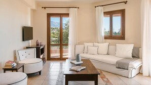 A living room with a sofa, a coffee table, a chair, and a television. There are curtains and a view of the outdoors through a glass door. - Ilios Kefalonia With 3 Bedrooms and 3 Bathrooms (Kefalonia)